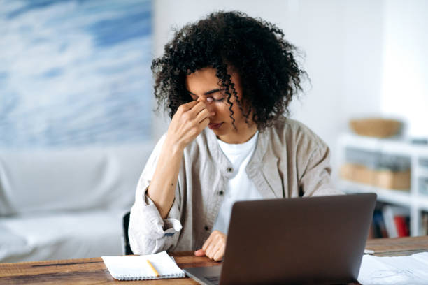 Tired frustrated overworked African American young woman in casual wear, manager, freelancer or student sitting in living room at laptop, experiencing stress, headache, rubs and close her eyes with stock photo