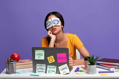 A Lot Of Work Concept. Portrait of tired female freelancer sleeping at workplace with stickers on eyes, sitting at table over purple studio background. Distracted woman at home office