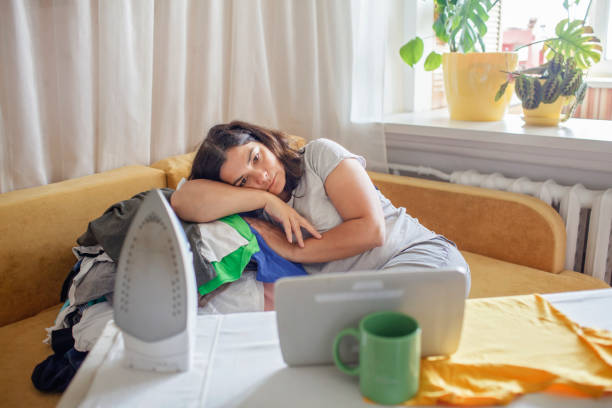 Tired middle aged woman watches video on tablet and ironing things, housewife and mom burnout stock photo