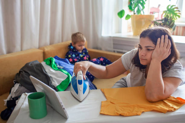 Tired mom watches video on tablet and ironing things, next to her son with smartphone too stock photo