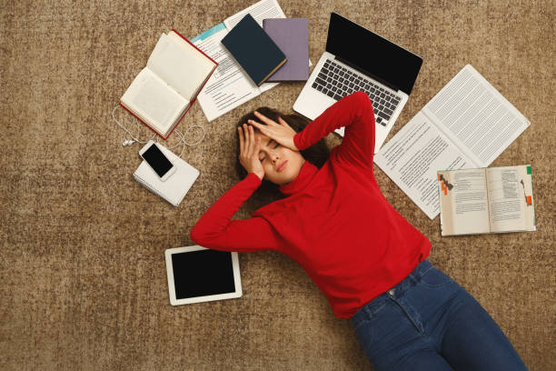 Tired student girl lying on the floor with books and gadgets Exhausted student girl lying on the floor among textbooks, tests and gadgets, copy space. Woman holding head with hands, got tired while preparing for exams. Education and overworking concept  student life stock pictures, royalty-free photos & images