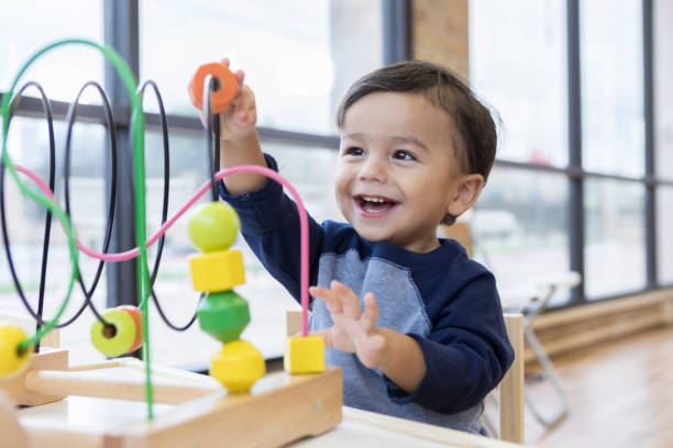 Toddler boy enjoys playing with toys in waiting room An adorable toddler boy sits at a table in a doctor's waiting room and reaches up cheerfully to play with a toy bead maze. preschool stock pictures, royalty-free photos & images