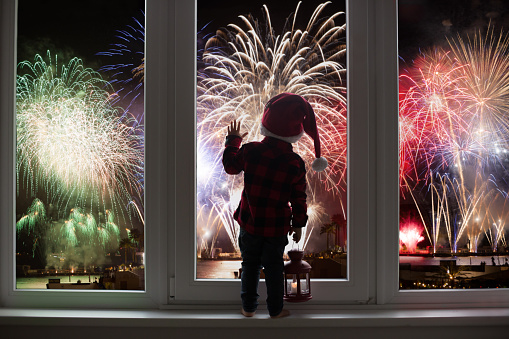 Toddler child standing in front of a big french doors, leaning against it looking at new years eve fireworks
