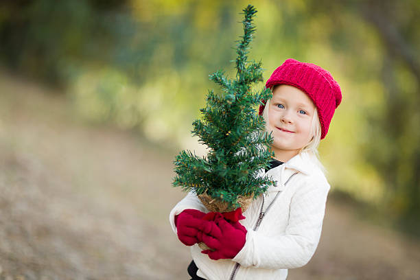 Toddler In Red Mittens and Cap Holding Small Christmas Tree Baby Girl In Red Mittens and Cap Holding Small Christmas Tree Outdoors. planting chrismas tree stock pictures, royalty-free photos & images