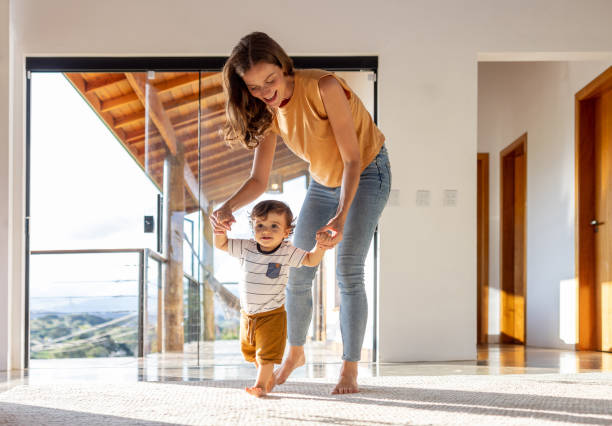 Toddler learning how to walk at home with the help of his mother stock photo