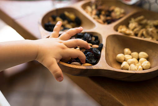 Child hand picking almonds from a wooden bowl with mixed nuts and dried fruits.