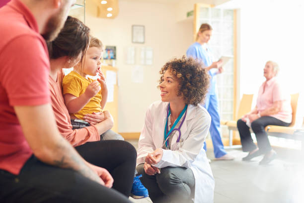 Toddler talking to female doctor A young family sits talking to the doctor. The toddler is sat on his mother’s knee as the doctor kneels down to talk to him general practitioner stock pictures, royalty-free photos & images