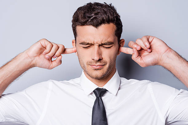 Too loud sound. Frustrated young man in shirt and tie holding fingers in ears and keeping eyes closed while standing against grey background Fingers in Ears stock pictures, royalty-free photos & images