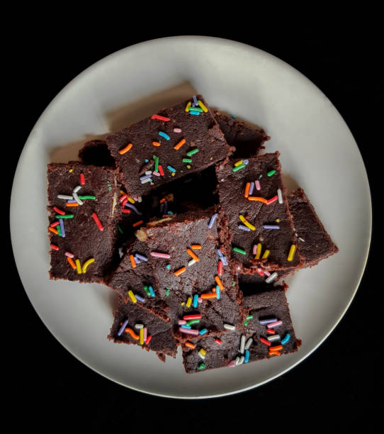 Top down angle photo of stacked pieces of Chocolate Biscuit Sprinkle Cakes arranged on a white plate and prepared pieces in the background on a kitchen top stock photo