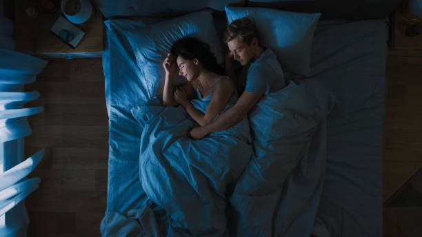 top view bed at night: attractive young couple sleeping together, holding each other in arms, embracing. blue nightly colors with cold weak lamppost light shining through the window. - sleeping couple imagens e fotografias de stock