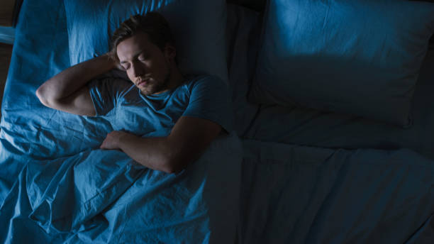 Top View of Handsome Young Man Sleeping Cozily on a Bed in His Bedroom at Night. Blue Nightly Colors with Cold Weak Lamppost Light Shining Through the Window. Top View of Handsome Young Man Sleeping Cozily on a Bed in His Bedroom at Night. Blue Nightly Colors with Cold Weak Lamppost Light Shining Through the Window. man sleeping in bed top view stock pictures, royalty-free photos & images