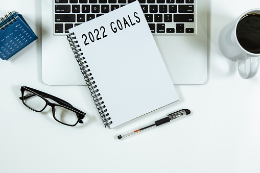 Top view of notebook handwriting goals for 2022 year, laptop and eyeglasses on white background. Business plan concept.
