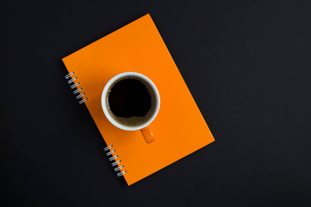 Top view of orange coffee cup on the orange notebook on the black background. Copy space. Close-up. stock photo
