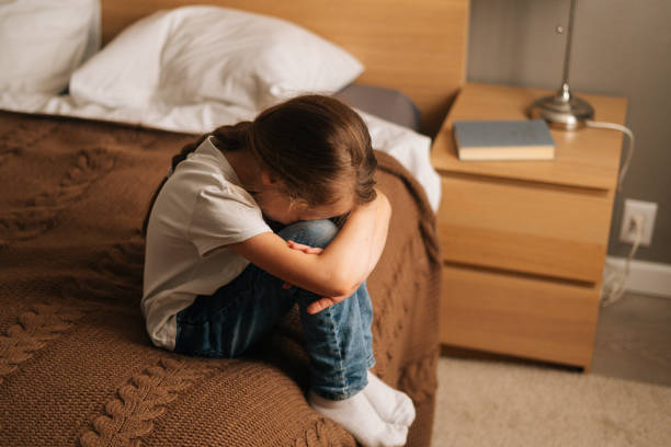 Top view of upset little girl hugging knee, sobbing with head bowed and crying sitting alone on bed in bedroom. stock photo