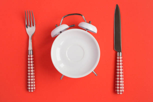 Top view of white saucer on the dial of the alarm clock, knife and fork on the red background. Copy space. stock photo