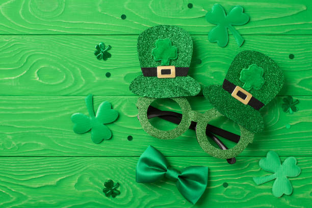 top view photo of st patrick's day decorations shamrocks trefoil shaped confetti funny outfit hat shaped party glasses and green bow-tie on isolated textured green wooden desk background - carnival accessories flat lay imagens e fotografias de stock