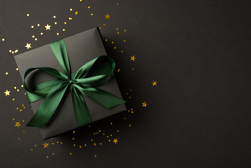 Top view photo of stylish giftbox with green ribbon bow golden stars and confetti on isolated black background with copyspace