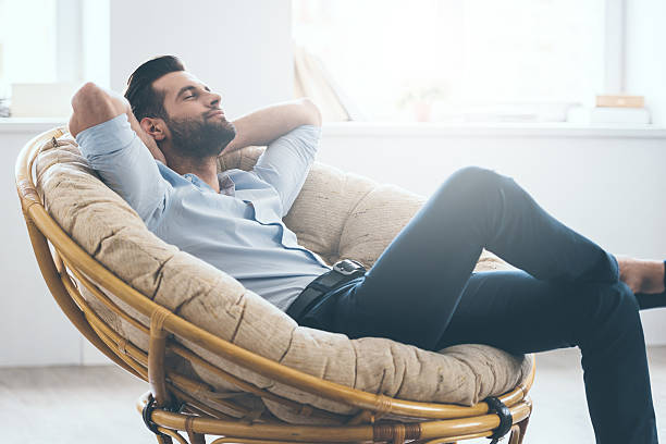 Total relaxation. Handsome young man keeping eyes closed and holding hands behind head while sitting in big comfortable chair at home day dreaming photos stock pictures, royalty-free photos & images
