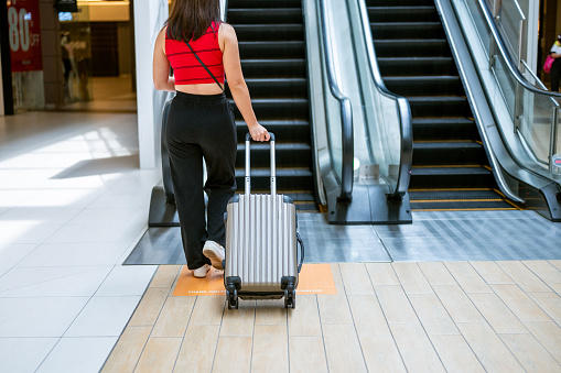 Young Asian female rides the escalator up at the airport. She is holding her luggage handle and passport ready to embark on a new travel adventure.