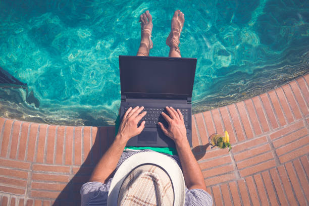 Tourist sitting at the edge of swimming pool and using blank screen laptop. Summer vacation and freelancer concepts stock photo