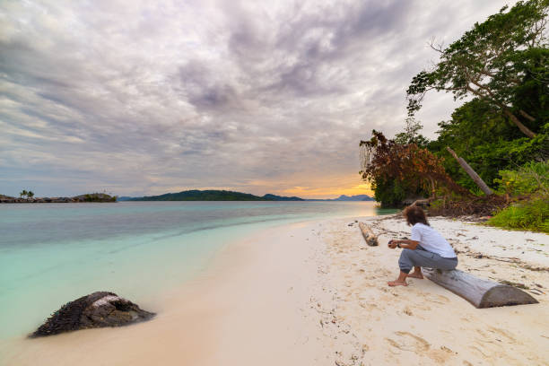 Tourist watching a relaxing sunset sitting on the beach in the remote Togean Islands, Central Sulawesi, Indonesia, upgrowing travel destination in recent years. Wide angle view. Tourist watching a relaxing sunset sitting on the beach in the remote Togean Islands, Central Sulawesi, Indonesia, upgrowing travel destination in recent years. Wide angle view. desert island stock pictures, royalty-free photos & images
