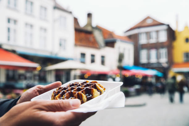 Traditional Belgian dessert, pastry. Tourist holds in hand popular street food - Belgium tasty waffle with chocolate sauce on the background of city tourist streets of Bruges, Belgium, Europe. Traditional Belgian dessert, pastry. brugge, belgium stock pictures, royalty-free photos & images