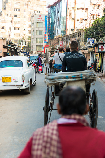 Calcutta, India - January 4, 2014: Urban street, some rickshaws with tourists in line driving on the street, a car at the side, some people walking, high houses in background. Photo taken from the rickshaw. 