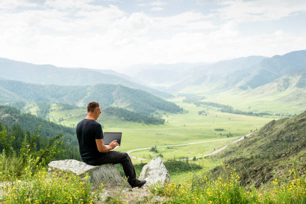 Traveler blogger work remote on netbook computer while enjoying mountain nature landscape view outdoors. man working outdoors with laptop. stock photo