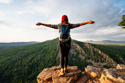 A traveler girl with a backpack is standing on the edge of the mountain, a rear view. A young woman with a backpack standing on the edge of a cliff and looking at the sky with her hands raised.