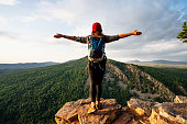 istock A traveler girl with a backpack is standing on the edge of the mountain, a rear view. A young woman with a backpack standing on the edge of a cliff and looking at the sky with her hands raised. 1334470825
