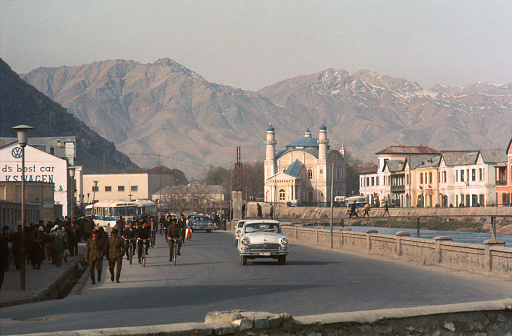 Kabul, 1969,  afternoon commuters make their way beside the river Darya-e-Kabul, by car, by bus, by bicycle and on foot.  On the opposite bank of the river, stands Shah Du Shamshira Mosque and beyond the snow topped mountains that surround the city. The river is crossed by a narrow footbridge, now no longer there.  Recorded on Ektachrome - the date created is approximate.