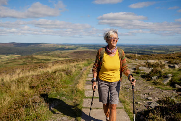 Travelling Over the Hills A senior woman walking on a footpath along the top of a hill using hiking poles with the landscape surrounding her in Rothbury, Northumberland. She is walking while smiling and looking ahead. rothbury northumberland stock pictures, royalty-free photos & images