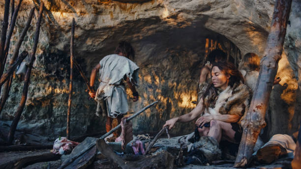 tribe of hunter-gatherers wearing animal skin live in a cave. leader brings animal prey from hunting, female cooks food on bonfire, girl drawing on wals creating art. neanderthal homo sapiens family - fire caveman imagens e fotografias de stock