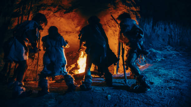 tribe of prehistoric hunter-gatherers wearing animal skins dance around bonfire outside of cave at night. neanderthal / homo sapiens family doing pagan religion dancing near fire back view slow motion - fire caveman imagens e fotografias de stock