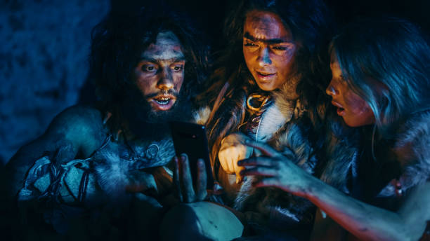 tribe of prehistoric, primitive hunter-gatherers wearing animal skins use smartphone in a cave at night. neanderthal / homo sapiens family browsing internet on mobile phone - fire caveman imagens e fotografias de stock