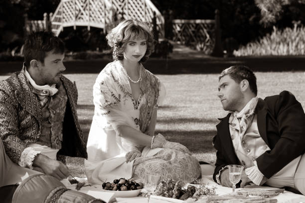 Trio of people dressed in vintage clothing on picnic blanket. Gentlemen stare off as beautiful lady looks to camera Trio of young people dressed in vintage clothing on picnic blanket. The gentlemen stare off as beautiful lady looks to camera romance book cover stock pictures, royalty-free photos & images