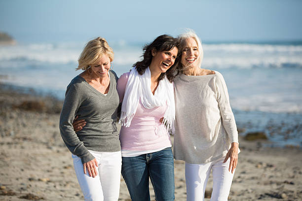 Trio of women walking at the beach Three smiling mature women embracing one another on a rocky, seaweed covered sandy beach.  The ocean and clear blue skies are behind them. 55 59 years stock pictures, royalty-free photos & images