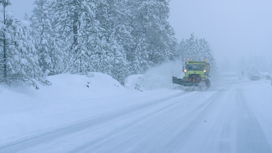 CLOSE UP: Truck plows the snowy country road during a horrible snowstorm raging in the state of Washington. Cars carefully drive behind a snow plough clearing the road during a devastating blizzard.
