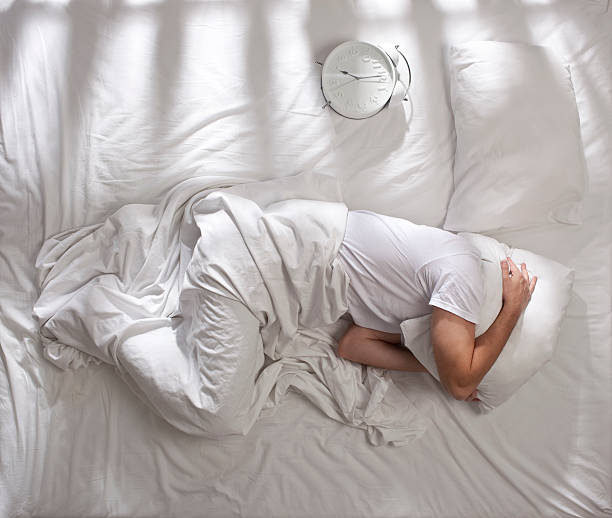 Trying to sleep. Man under white sheets and pillow on head trying to sleep in bed. White clock showing seven o'clock in the morning. Top view image. man sleeping in bed top view stock pictures, royalty-free photos & images