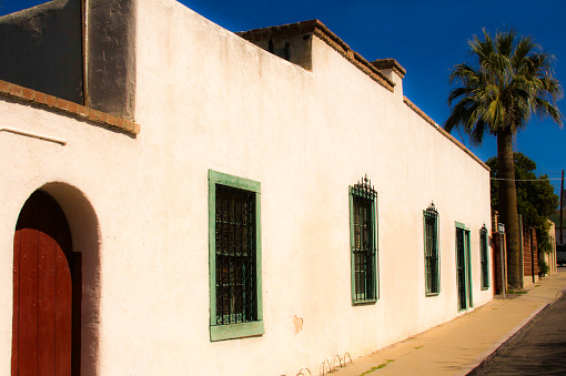 A street scene with a sunlit traditional white building, sidewalk, and palm tree in Tucson, AZ. Brilliant blue sky.
