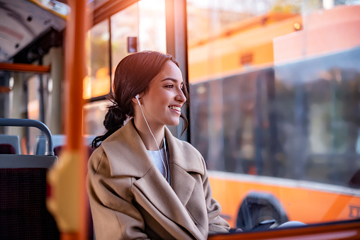 One Young Woman Smiling in a Bus While Listening to Music using Earphones. Beautiful Young Woman With Earphones Taking Bus to Work. The Passenger Listening to Music in the Bus or Train, Technology Lifestyle, Transportation and Traveling Concept.