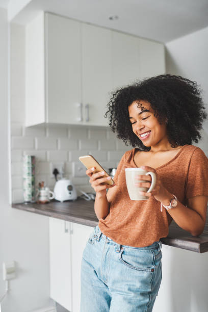 Turn on the day with coffee and social media Shot of a young woman using a smartphone and having coffee in the kitchen at home modern lifestyle stock pictures, royalty-free photos & images