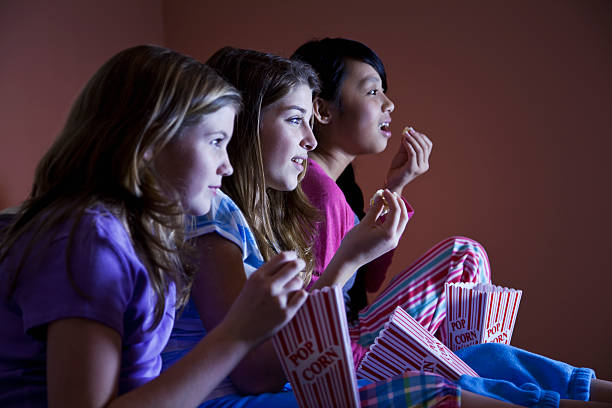 Tween girls watching TV Preteen girls (12 years) sitting on bed in pajamas, watching TV, eating popcorn.  Focus on girl in middle. asian kids watching tv stock pictures, royalty-free photos & images