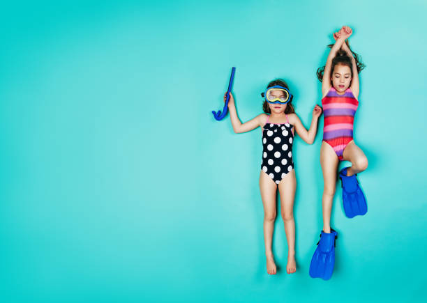 Twin girls pretending to be snorkelling Two girls lying down with snorkelers and flippers. Top view of twin girls wearing swimwear on blue background. little girls in bathing suits stock pictures, royalty-free photos & images