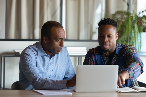 Two African American employees working on project together, using laptop pointing at screen, discussing strategy and sharing ideas at meeting, colleagues involved in teamwork, cooperation concept