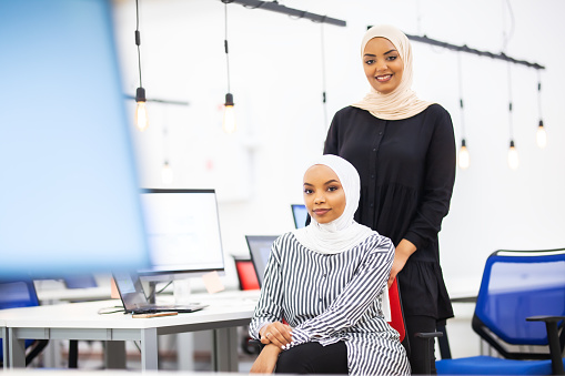 Two african american muslim girl colleagues with hijab posing together in a modern office. Monitors and light bulbs in the background and foreground
