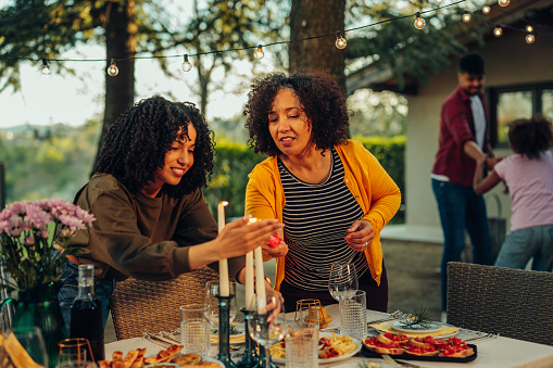 Two smiling biracial women arranging dining table and lighting candle on outdoors dining table, while man and daughter playing around