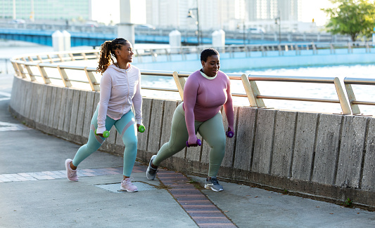 Two African-American woman exercising together in a city park, by a large fountain on a city waterfront. They are wearing long sleeved sports clothing. The woman with short hair in the dark pink shirt is a plus size model with a large build. She is in her 30s and her friend is in her 40s. They are doing lunges, carrying handweights.