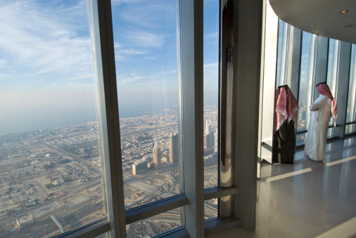 Two arab men in traditional dress looking out of the window over Dubai from the Burj Khalifa, the tallest building on earth.