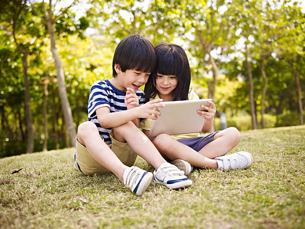 two asian children using tablet outdoors little asian girl and boy sitting on grass using digital tablet outdoors in a park. child korea little girls korean ethnicity stock pictures, royalty-free photos & images